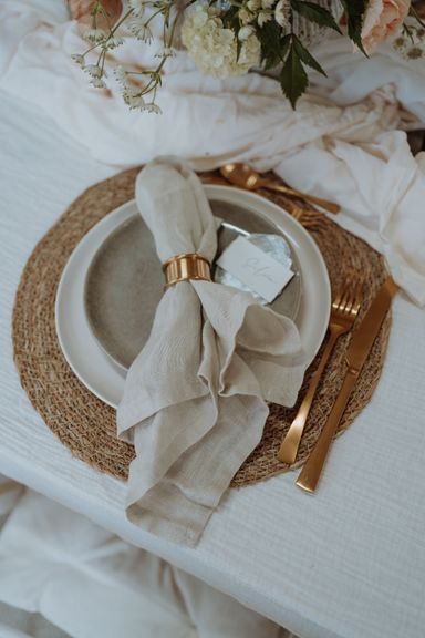 Detailaufnahme einer Tischdekoration, bei der man eine beige Leinenserviette mit goldenem Serviettenring auf einem Teller und Tischset sieht